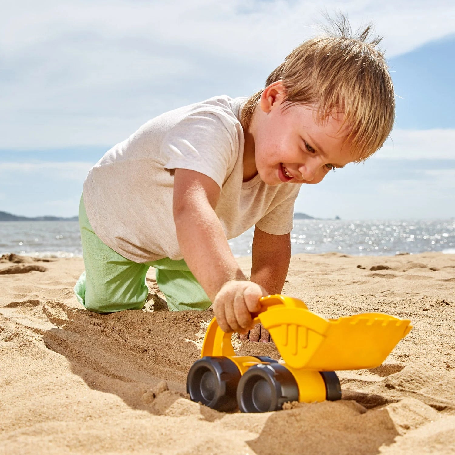A child playing with a Hape Monster Digger in the sand. The child is using the digger to scoop up sand and pile it up. The digger is yellow and green with a large, scoop-shaped bucket on the front.